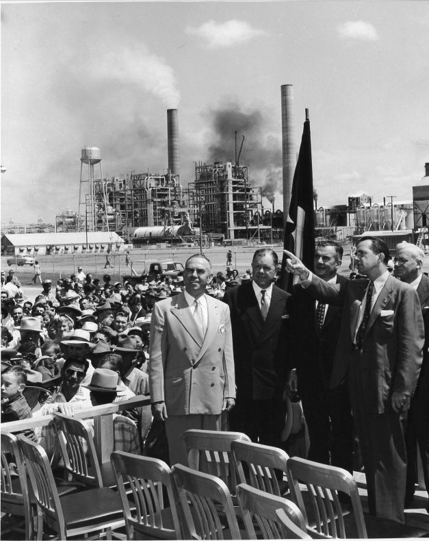 Texas dignitaries standing in front of the crowd at the Rockdale plant dedication, with the smelter and smokestacks behind them