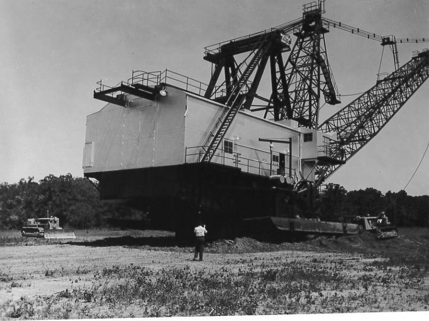A giant walking dragline excavator at a lignite mine, with a worker and bulldozers dwarfed beside it