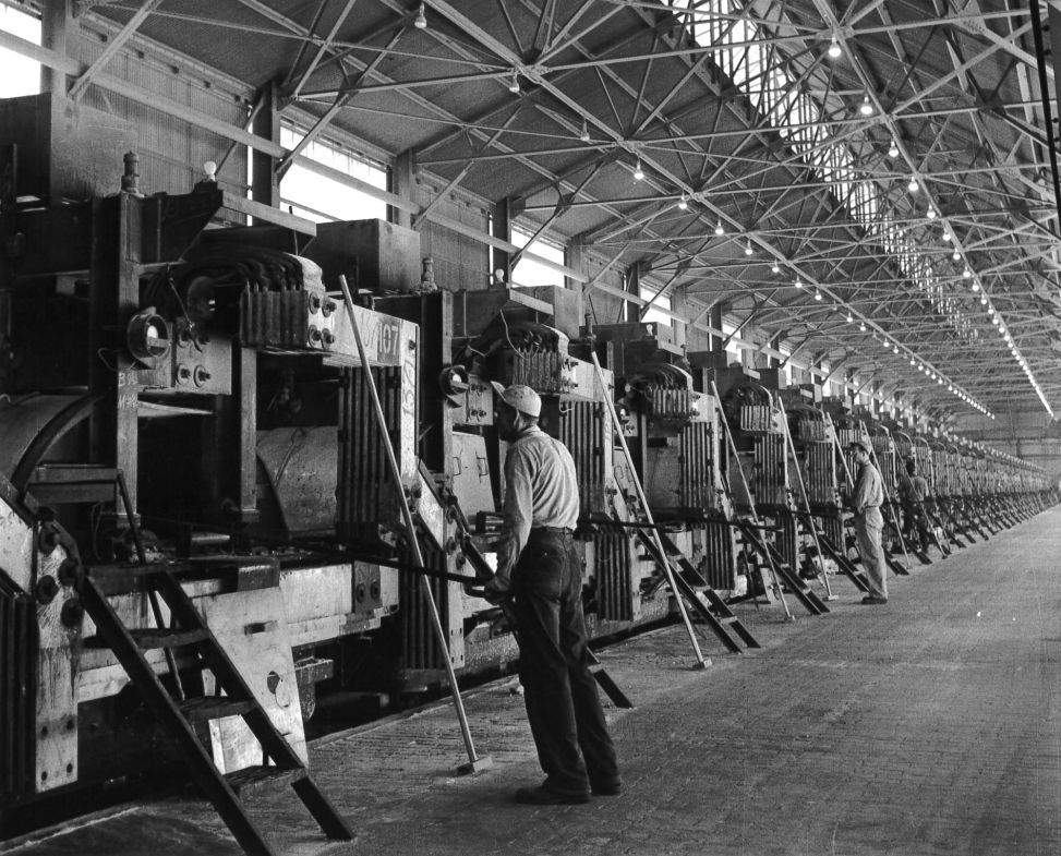 Workers tending a long line of aluminum reduction pots inside a smelting building